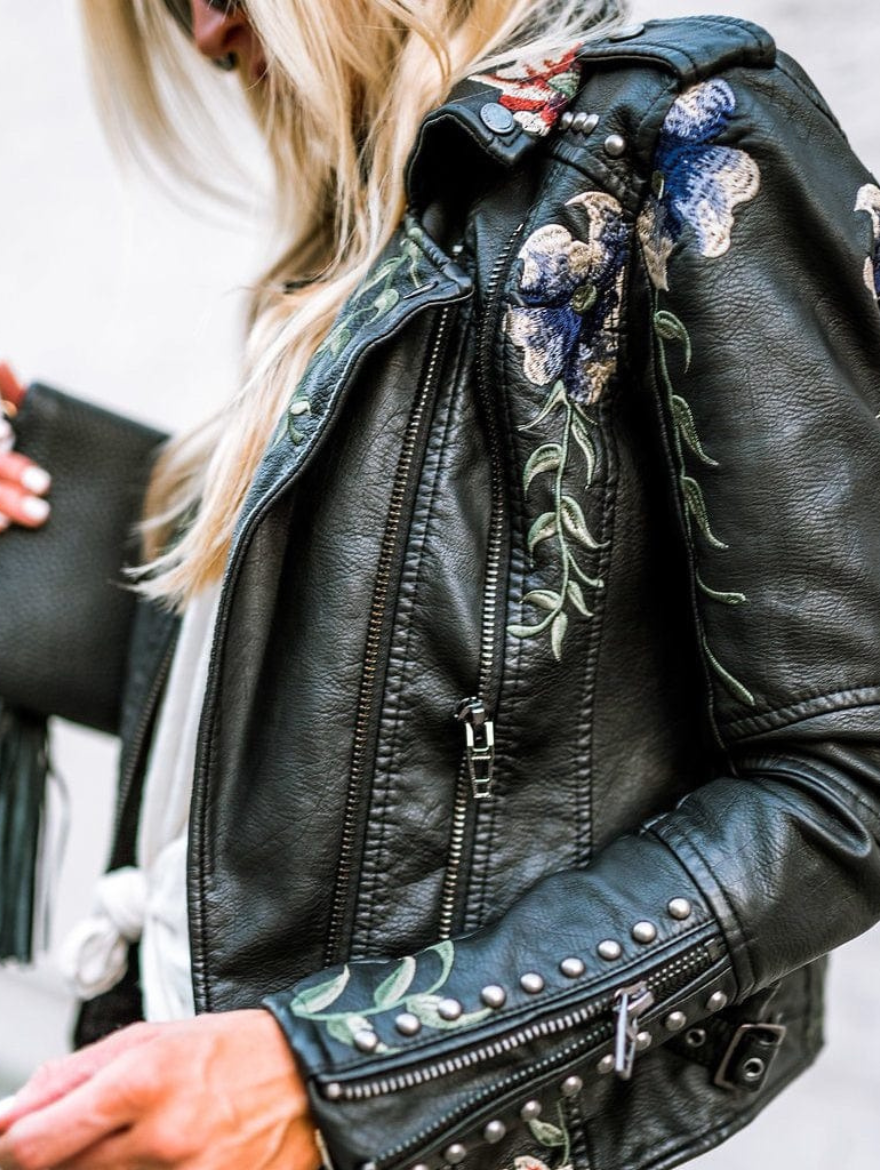 Veste en cuir noir, zips multiples et broderies florales bleues et vertes sur l’épaule.