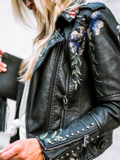 Veste en cuir noir, zips multiples et broderies florales bleues et vertes sur l’épaule.