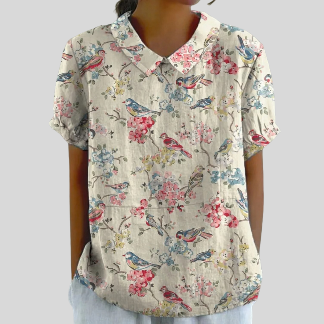 Blouse femme beige à manches courtes, col boutonné, motif oiseaux et fleurs multicolores.